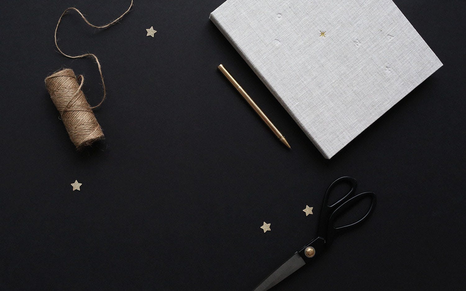 Tråd, sax och anteckningsblock med penna på svart underlag omgiven av små dekorationsstjärnor
