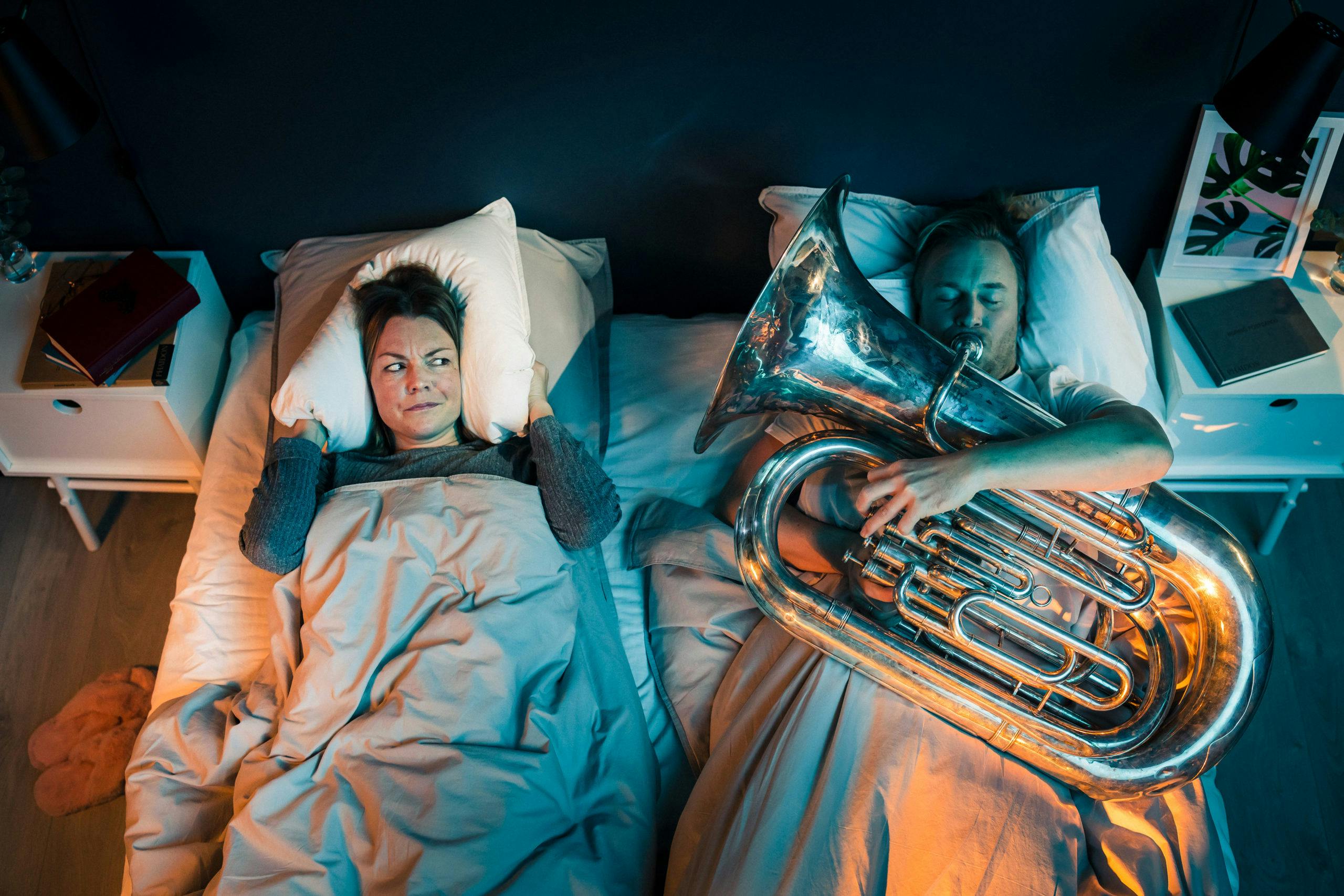 Man med trombon i sängen bredvid irriterad kvinna som försöker sova