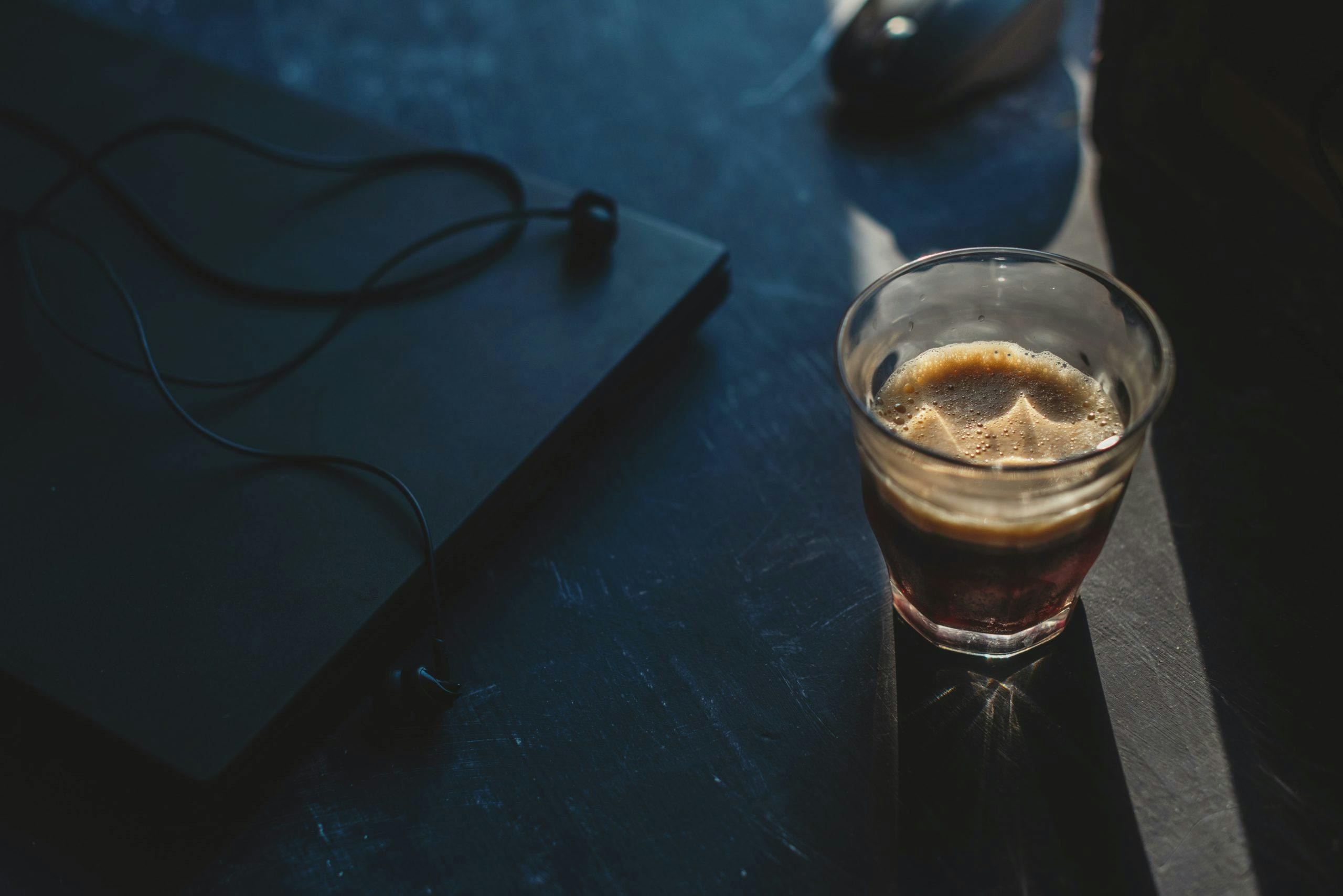 Glas med kaffe bredvid stängd laptop och hörlurar på bord