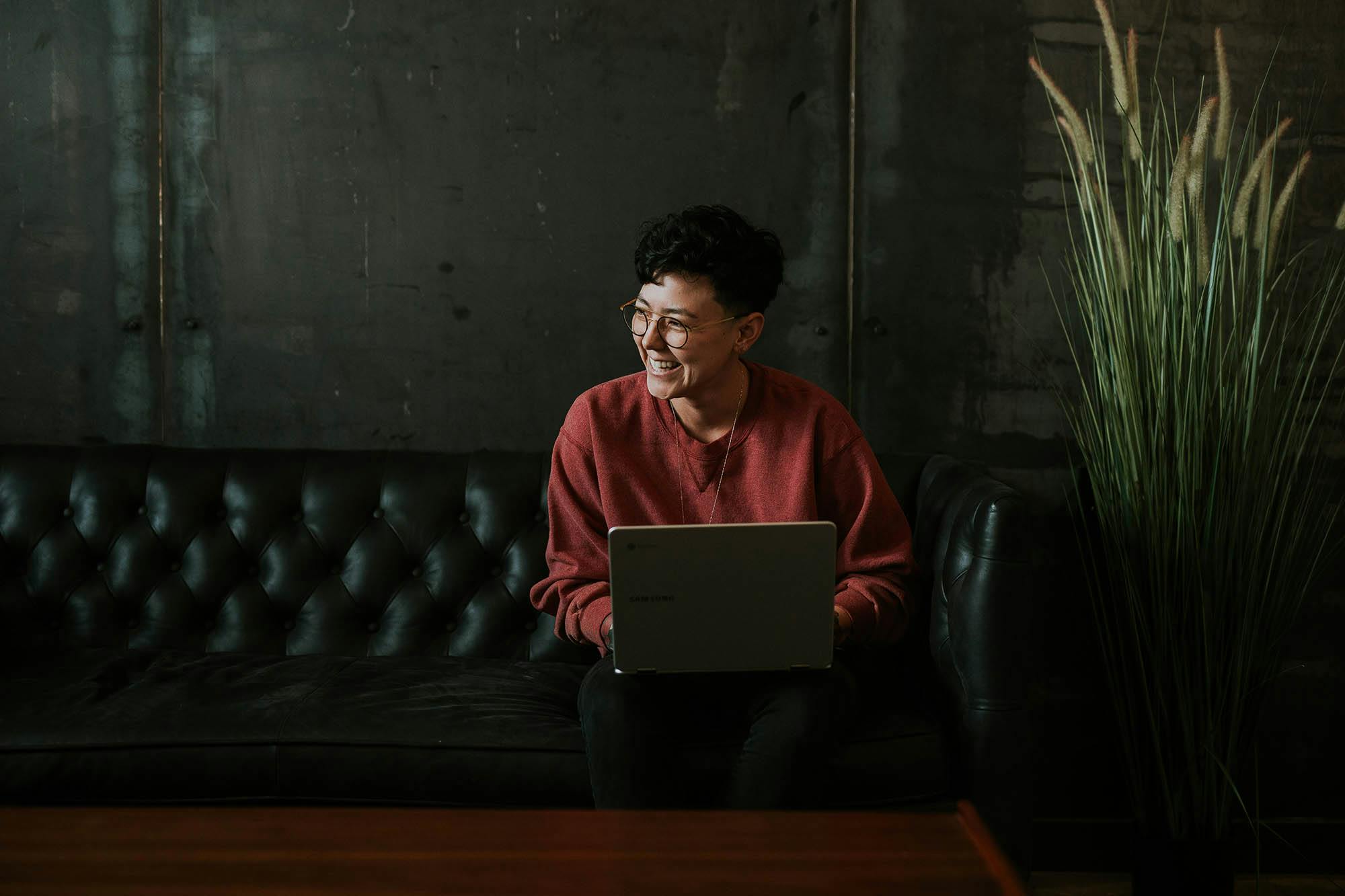 Kvinna skrattar med en laptop i sitt knä sittandes i en soffa